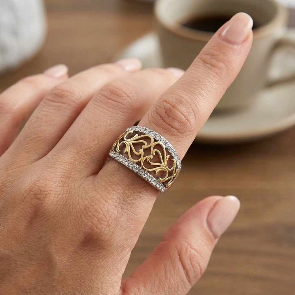 Gold ring with intricate design on a finger, blurred coffee cup in the background
